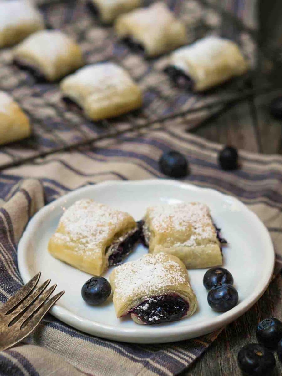 Angled view of 3 golden brown blueberry filled puff pastry rolls on a white plate - Hostess At Heart