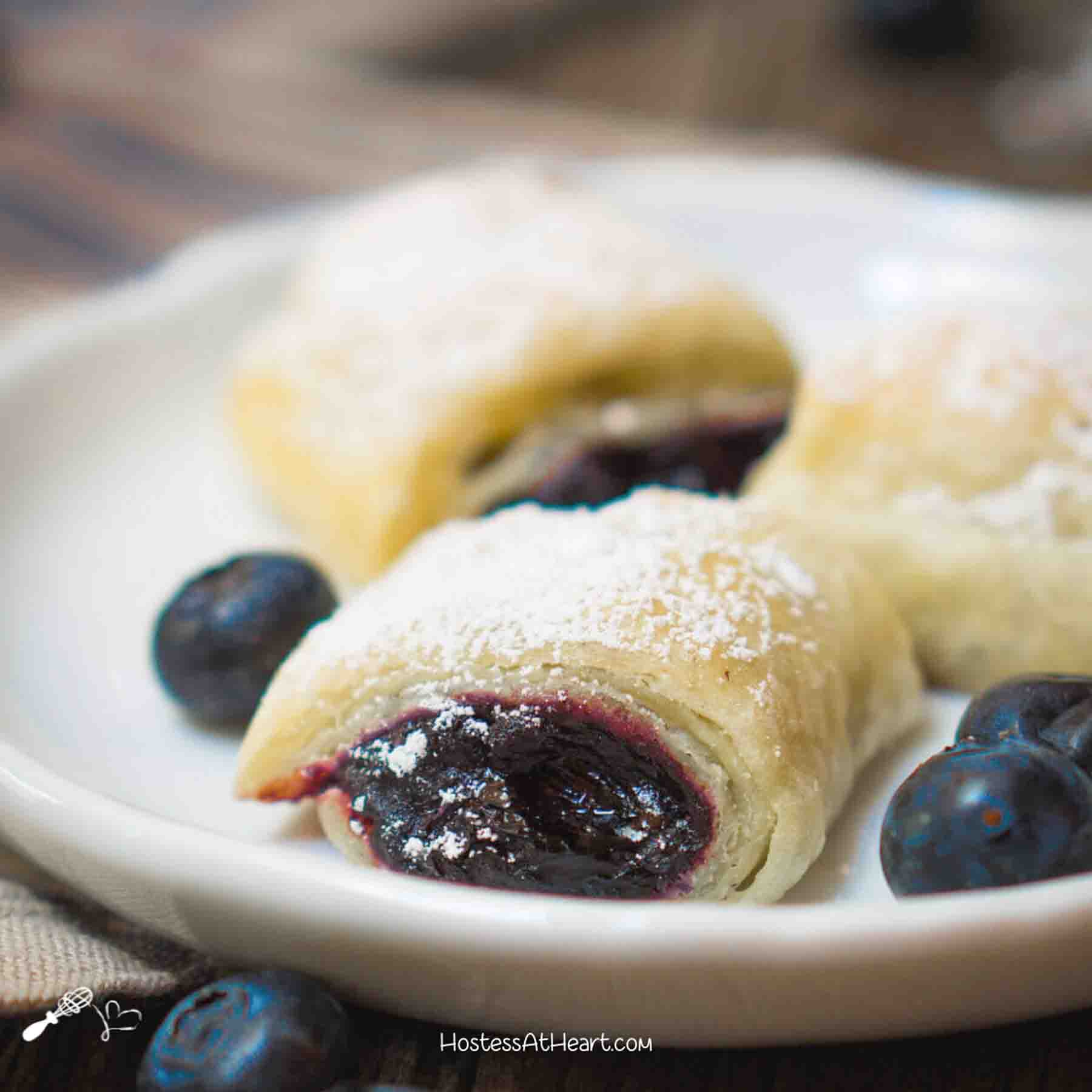 A white plate filled with blueberry puff pastry rolls dusted with powdered sugar - Hostess At Heart