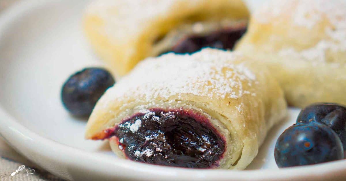 Sideview of a white plate topped with golden brown puff pastry blueberry rolls dusted with powdered sugar. Hostess At Heart
