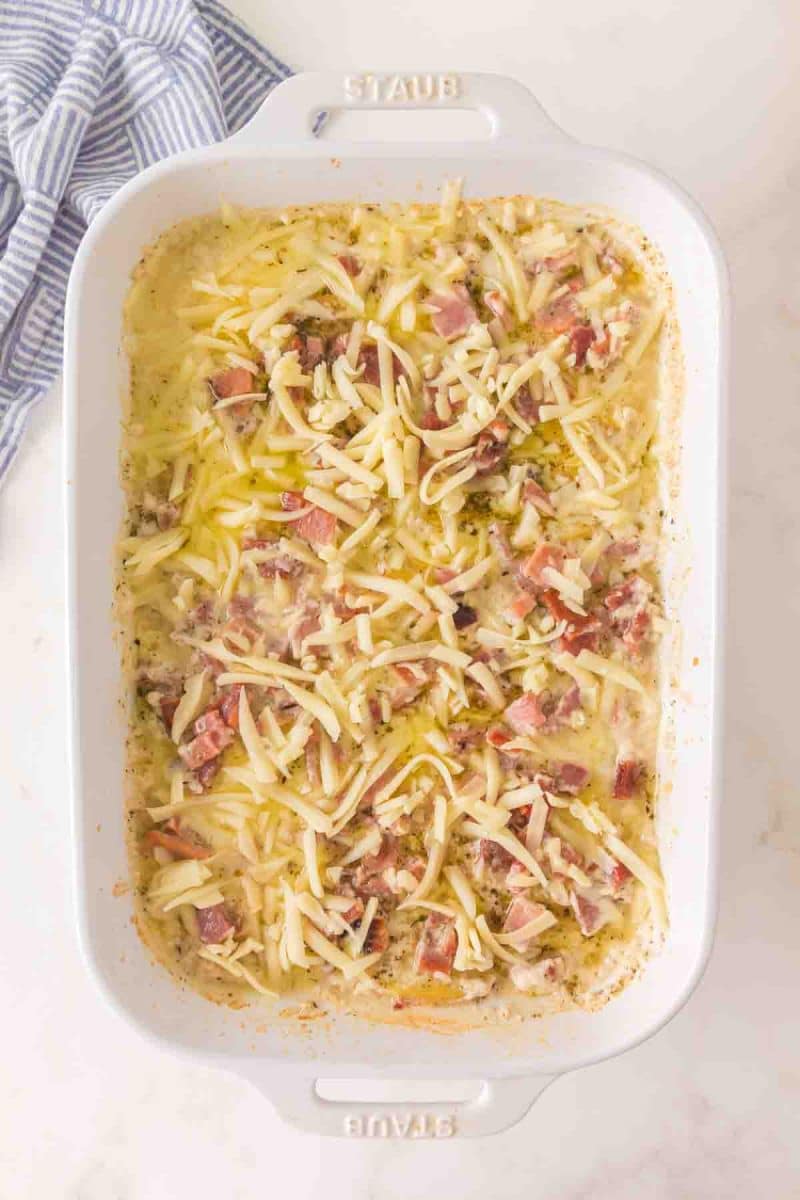Creamy ham and cheese sauce being poured over layer of sliced potatoes in 9x13 baking dish sprinkled with shredded cheese ready for baking - Hostess At Heart