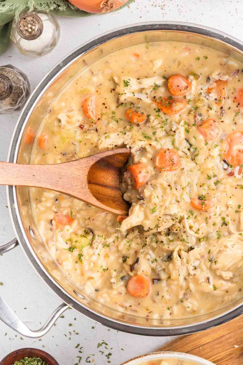 A wooden spoon dipped into a pot of creamy chicken and wild rice soup showing chunks of chicken, wild rice, and colorful vegetables in thick broth. Hostess At Heart