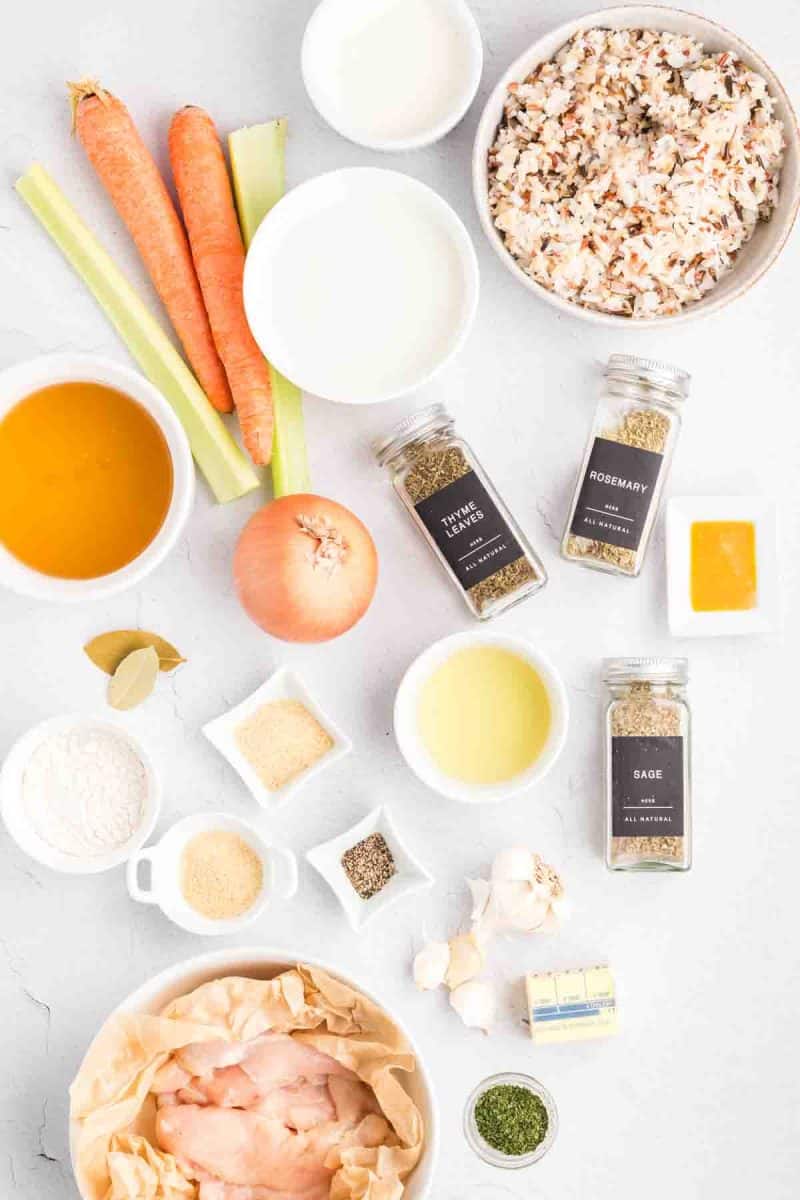 Raw ingredients for chicken and wild rice soup including wild rice blend, chicken tenders, fresh vegetables, and seasonings arranged on a white background. Hostess At Heart