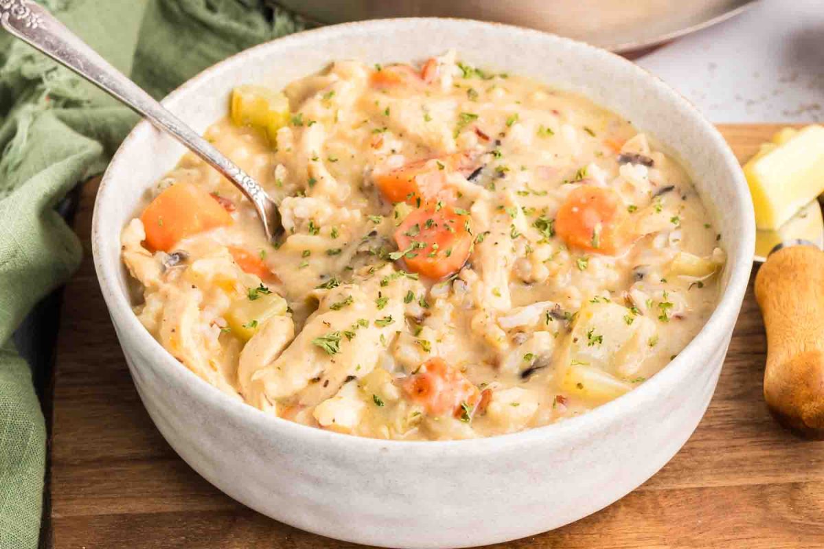 A spoon dipped into a bowl of creamy chicken and wild rice soup showing chunks of chicken, wild rice, and colorful vegetables in thick broth. Hostess At Heart