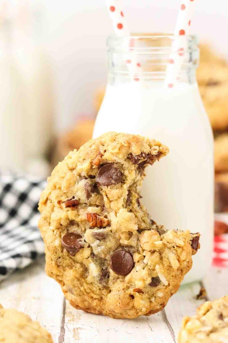 Extreme close-up of single cowboy cookie with a bite taken out of it sitting on it's end leaning against a milk bottle showing detailed texture with melted semi-sweet chocolate chips, golden toasted coconut flakes, oat pieces, and chopped pecans embedded throughout golden-brown cookie surface. Hostess At Heart