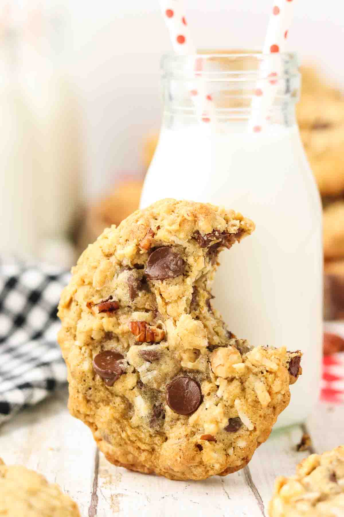 Extreme close-up of single cowboy cookie with a bite taken out of it sitting on it's end leaning against a milk bottle showing detailed texture with melted semi-sweet chocolate chips, golden toasted coconut flakes, oat pieces, and chopped pecans embedded throughout golden-brown cookie surface. Hostess At Heart