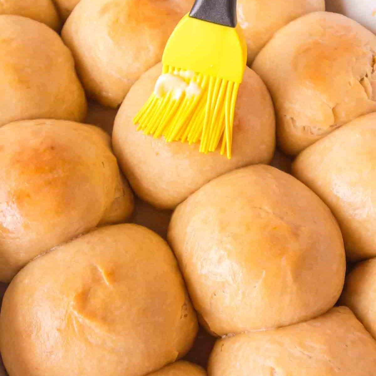 Close up of a basting brush applying butter to a pan of fluffy hawaiian rolls. Hostess At Heart