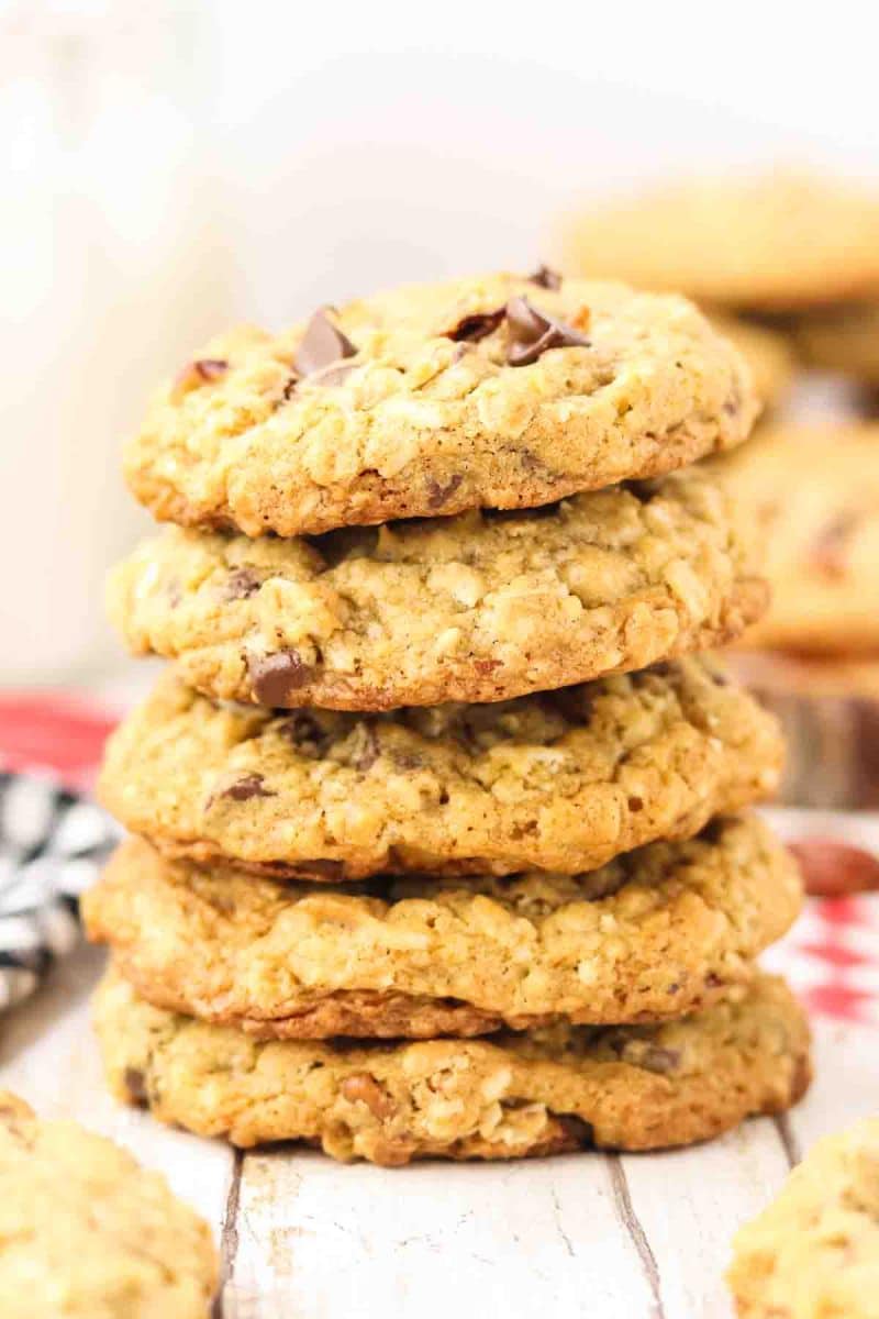 Tall stack of 5 golden-brown cowboy cookies on white wooden surface cookies showing rustic bumpy texture with visible chocolate chips melted on surface, toasted coconut strands, and pecan pieces. Hostess At Heart