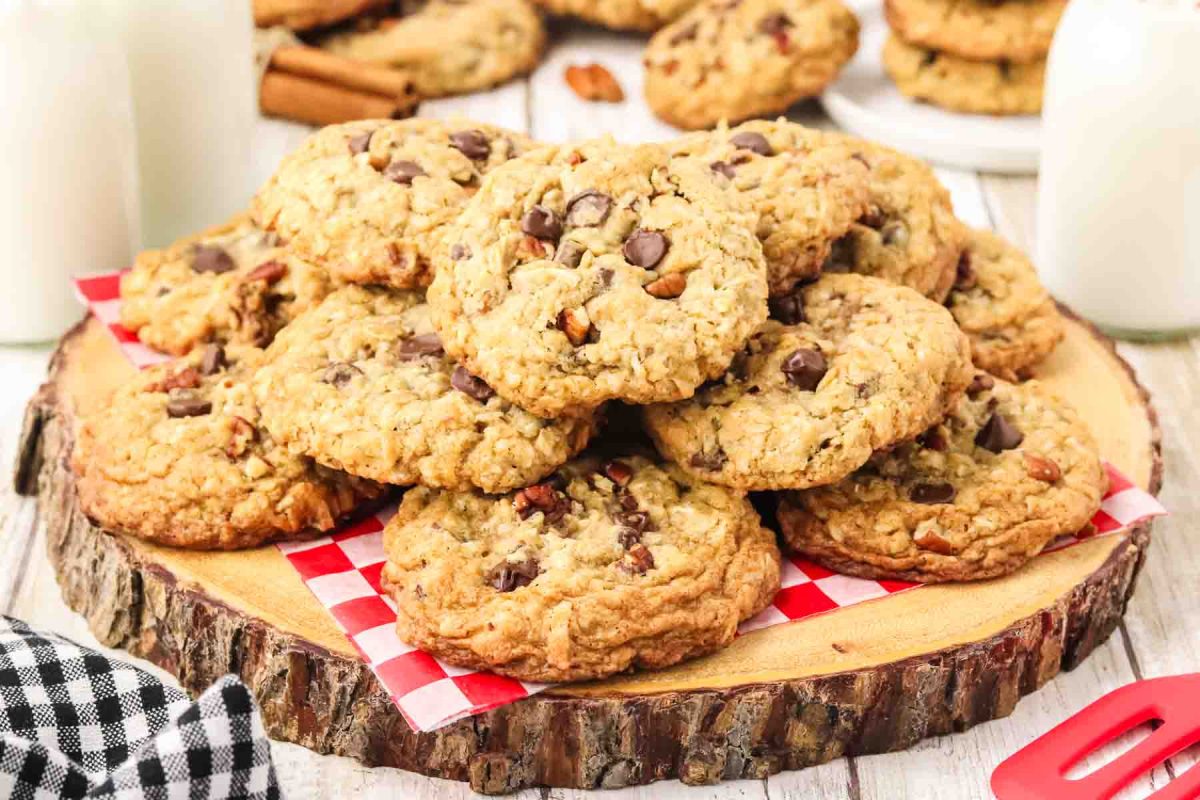 Sideiew of cowboy cookies piled on a wooden surface. The cookies have a soft bumpy texture and are loaded with chocolate chips, pecans and coconut. Hostess At Heart
