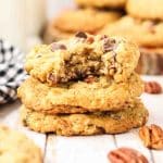 Stack of 3 golden-brown cowboy cookies on a wooden surface with a bite taken out of the top cookie. Cookies show rustic bumpy texture with visible chocolate chips melted on surface, toasted coconut strands, and pecan pieces. Hostess At Heart
