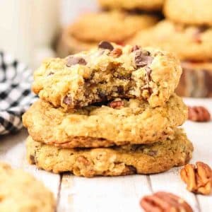 Stack of 3 golden-brown cowboy cookies on a wooden surface with a bite taken out of the top cookie. Cookies show rustic bumpy texture with visible chocolate chips melted on surface, toasted coconut strands, and pecan pieces. Hostess At Heart