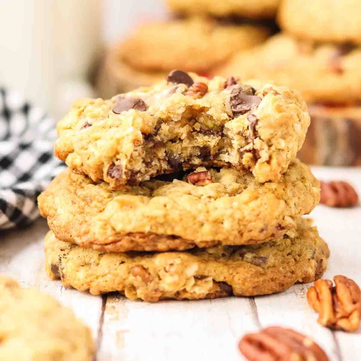 Stack of 3 golden-brown cowboy cookies on a wooden surface with a bite taken out of the top cookie. Cookies show rustic bumpy texture with visible chocolate chips melted on surface, toasted coconut strands, and pecan pieces. Hostess At Heart