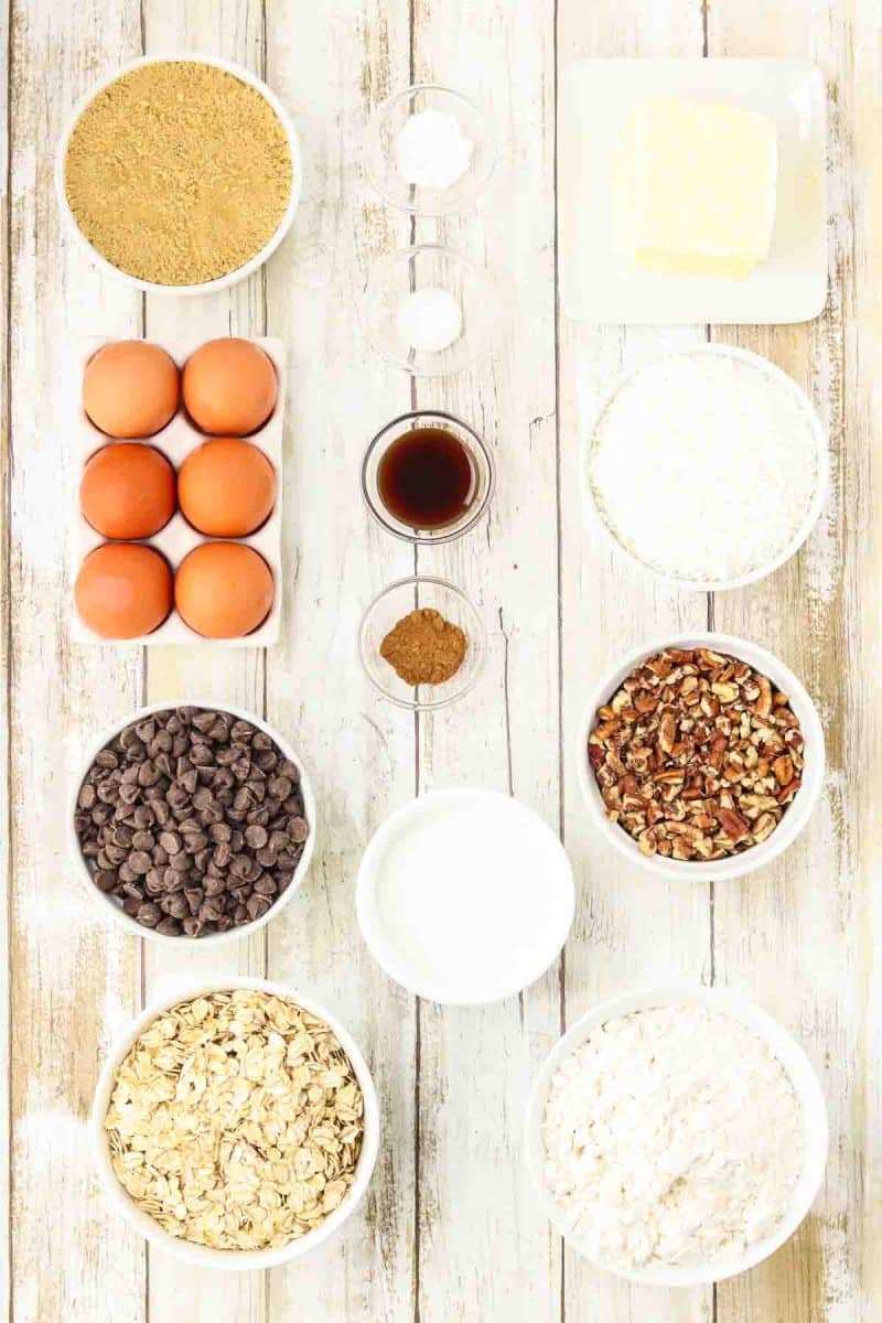 Cowboy cookie ingredients on a wooden background including flour, brown sugar, old-fashioned oats, shredded coconut, chocolate chips, chopped pecans, eggs, salt, baking soda, cinnamon, butter, and vanilla extract.