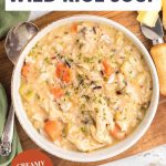 An image for photo of a bowl of creamy chicken and wild rice soup showing chunks of chicken, wild rice, and colorful vegetables in thick broth and garnished with herbs. Hostess At Heart