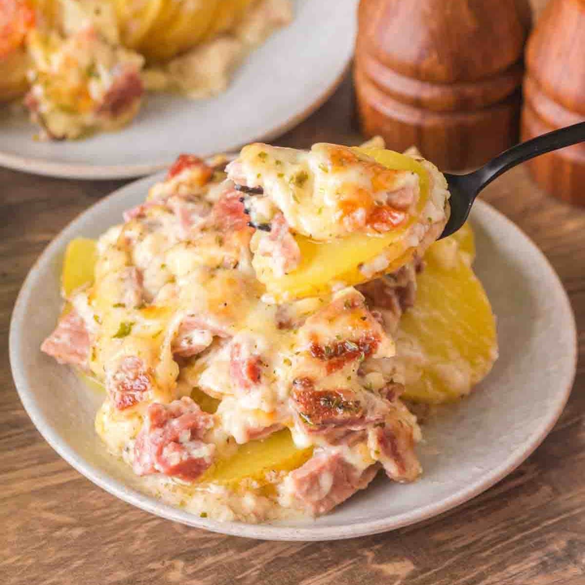 A fork lifting a bite of creamy scalloped potatoes with visible ham pieces and stretchy melted cheese from a plate - Hostess At Heart