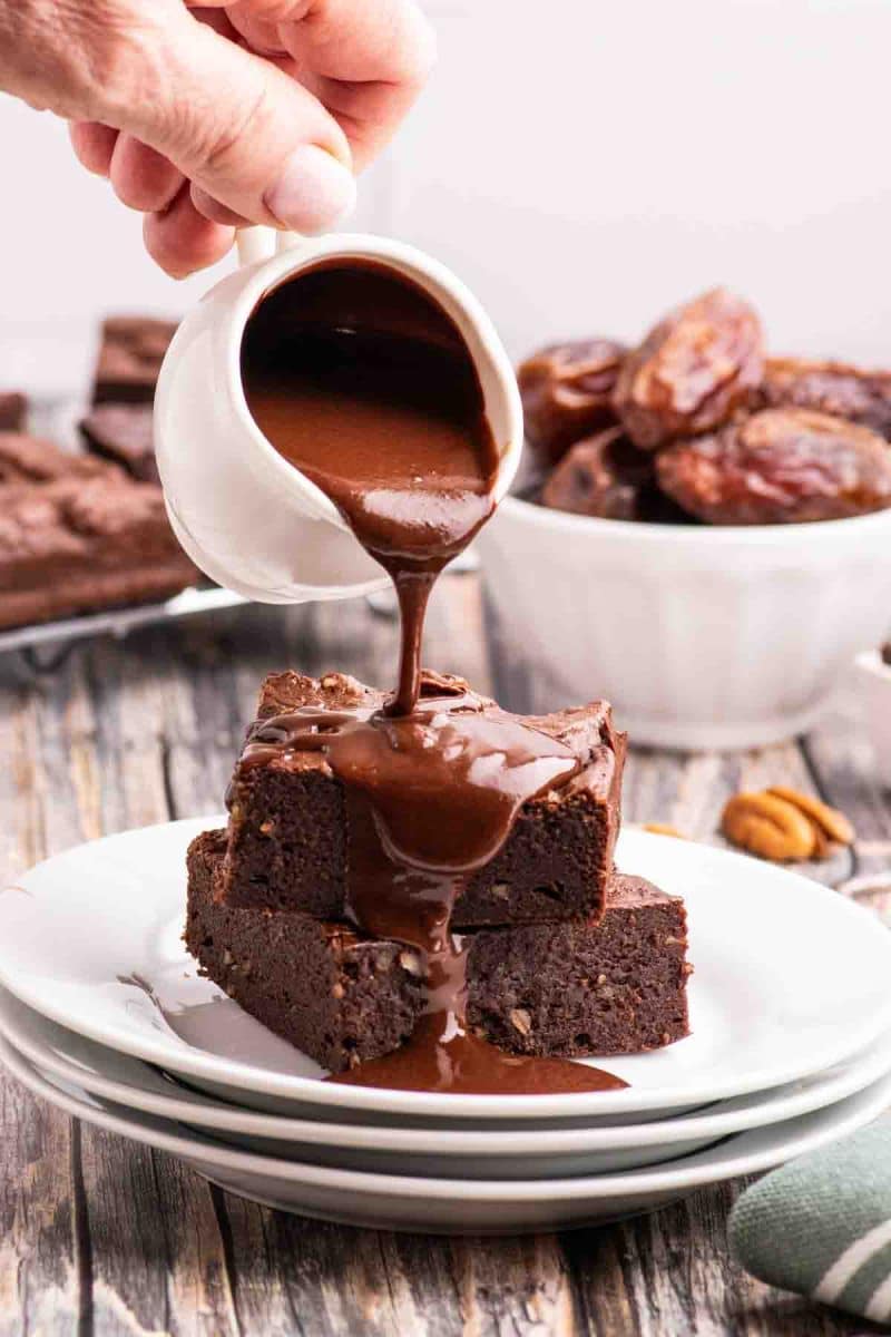 A white pitcher pouring chocolate ganache over a stack of two gluten-free brownies sitting on a white plate - Hostess At Heart