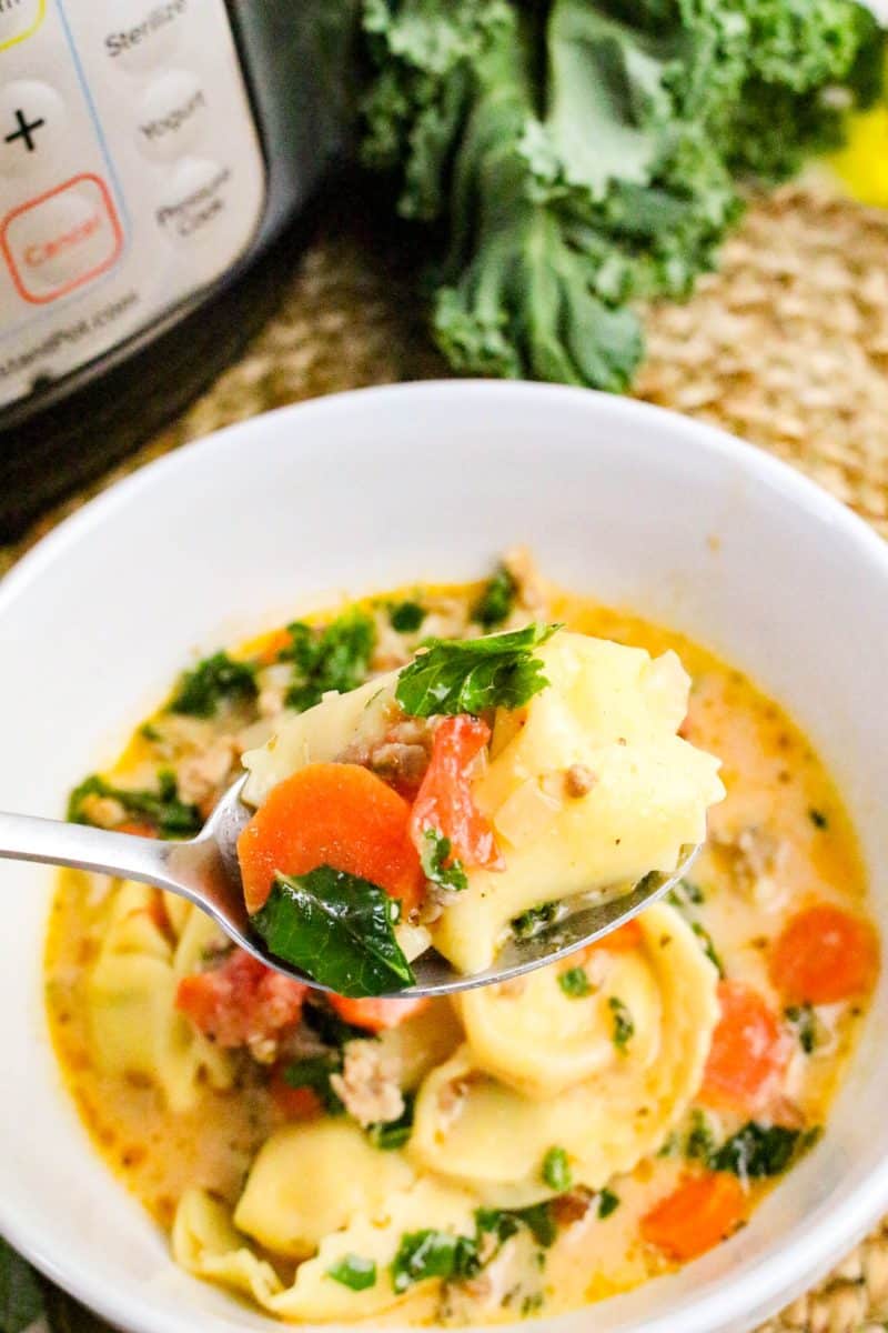 A spoon hovering over a white bowl filled with a bite of Cheese tortellini soup with Italian sausage, carrots, and chopped kale - Hostess At Heart