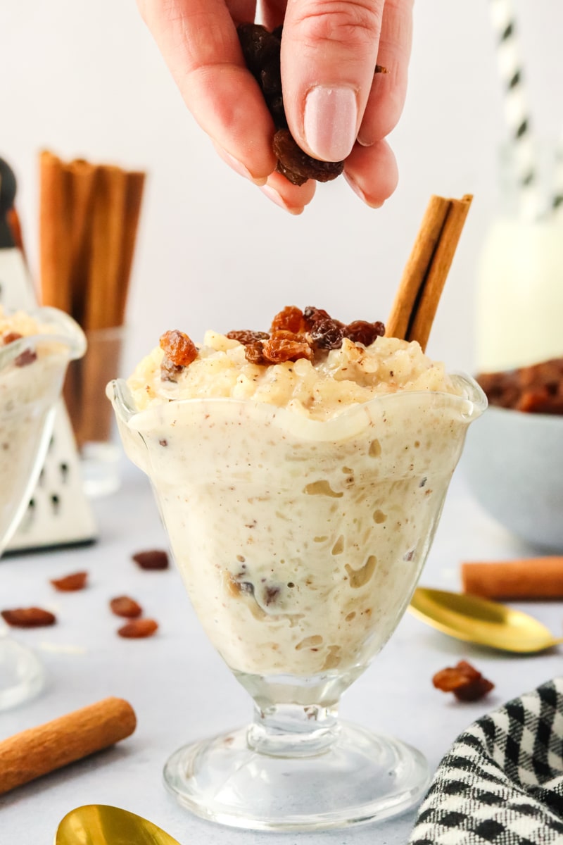 Sideview of a dessert cup filled with creamy instant pot rice pudding. A hand is sprinkling raisins over the top of the dessert - Hostess At Heart