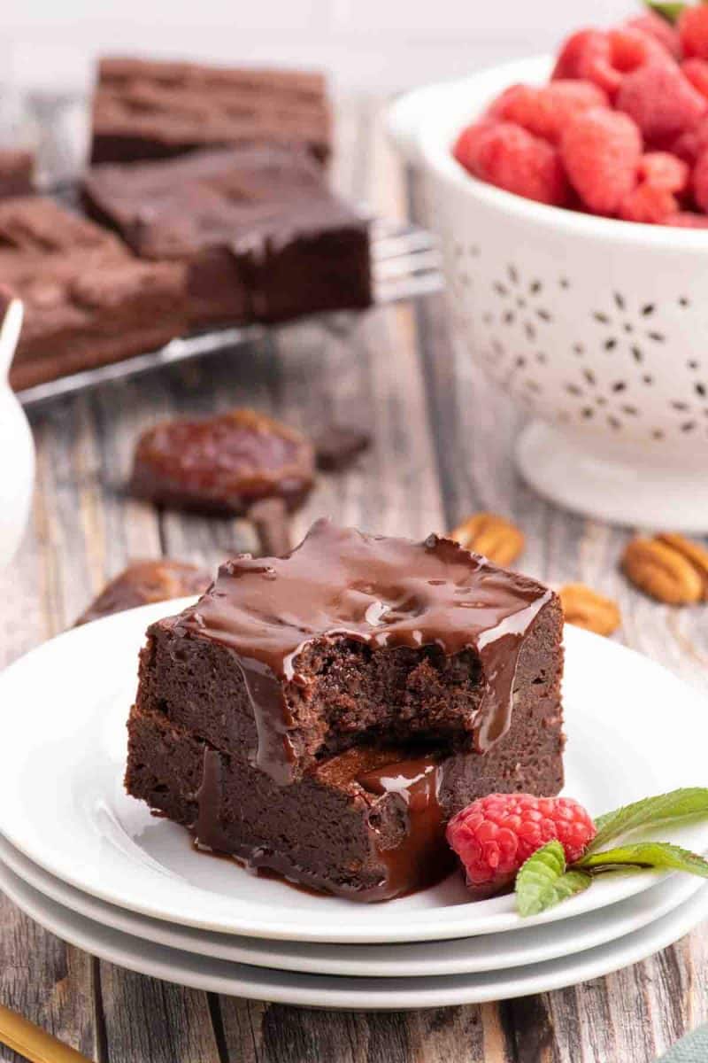 Sideview of two squares of cut brownies stacked on each other on a white plate. Dairy free chocolate ganache is poured over the top and running down the sides. The top brownie has a bite taken out of it showing a moist center. Hostess At Heart