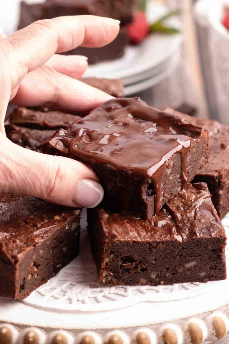 A hand lifting a square of date-sweetened chocolate brownies topped with dairy free granache - Hostess At Heart