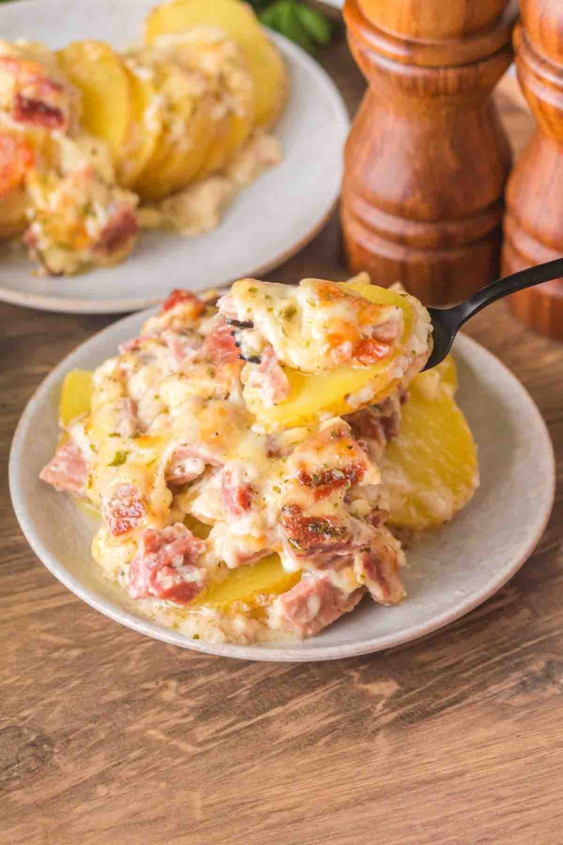 A table fork lifting a creamy bite of scalloped potatoes with visible ham pieces and stretchy melted cheese - Hostess At Heart