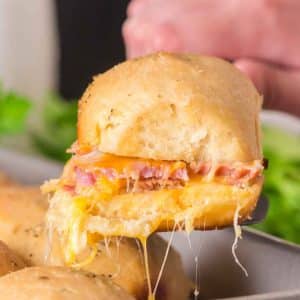 A spatula holding one Hawaiian ham and cheese slider from the pan showing stretchy melted cheese - Hostess At Heart