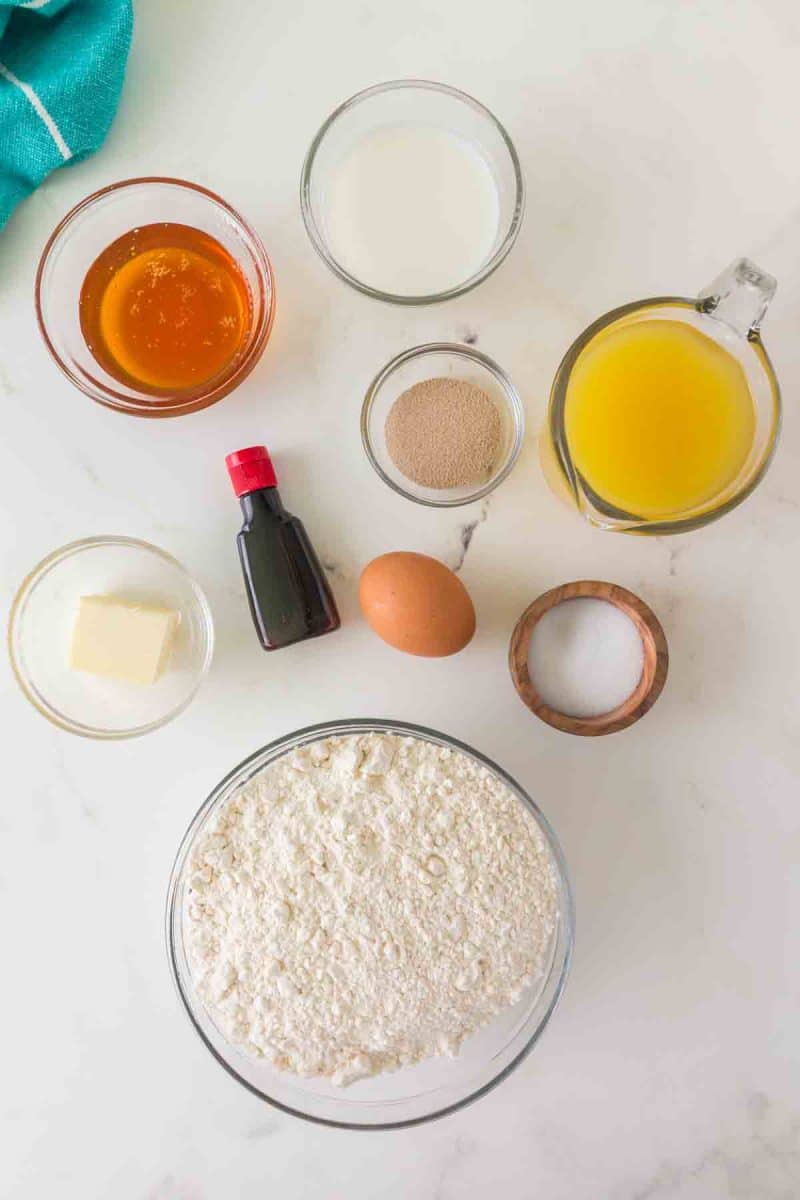 All ingredients for hawaiian rolls recipe arranged on marble counter including pineapple juice, buttermilk, salt, flour, yeast, eggs, butter, and honey - Hostess At Heart