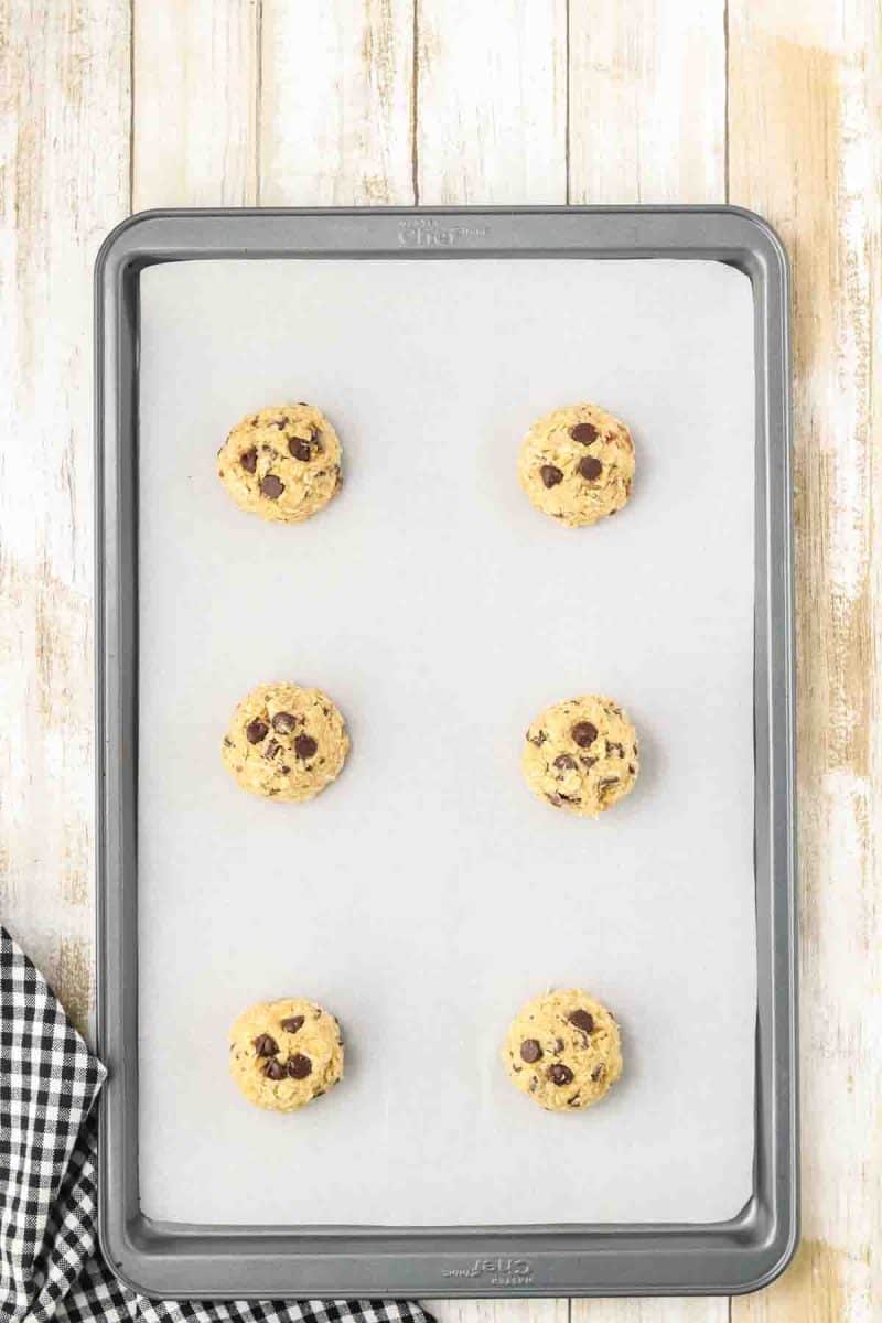 Six large rounded balls of cowboy cookie dough evenly spaced on parchment paper lined baking sheet ready for oven, each dough ball shows chunky texture with visible mix-ins. Hostess At Heart