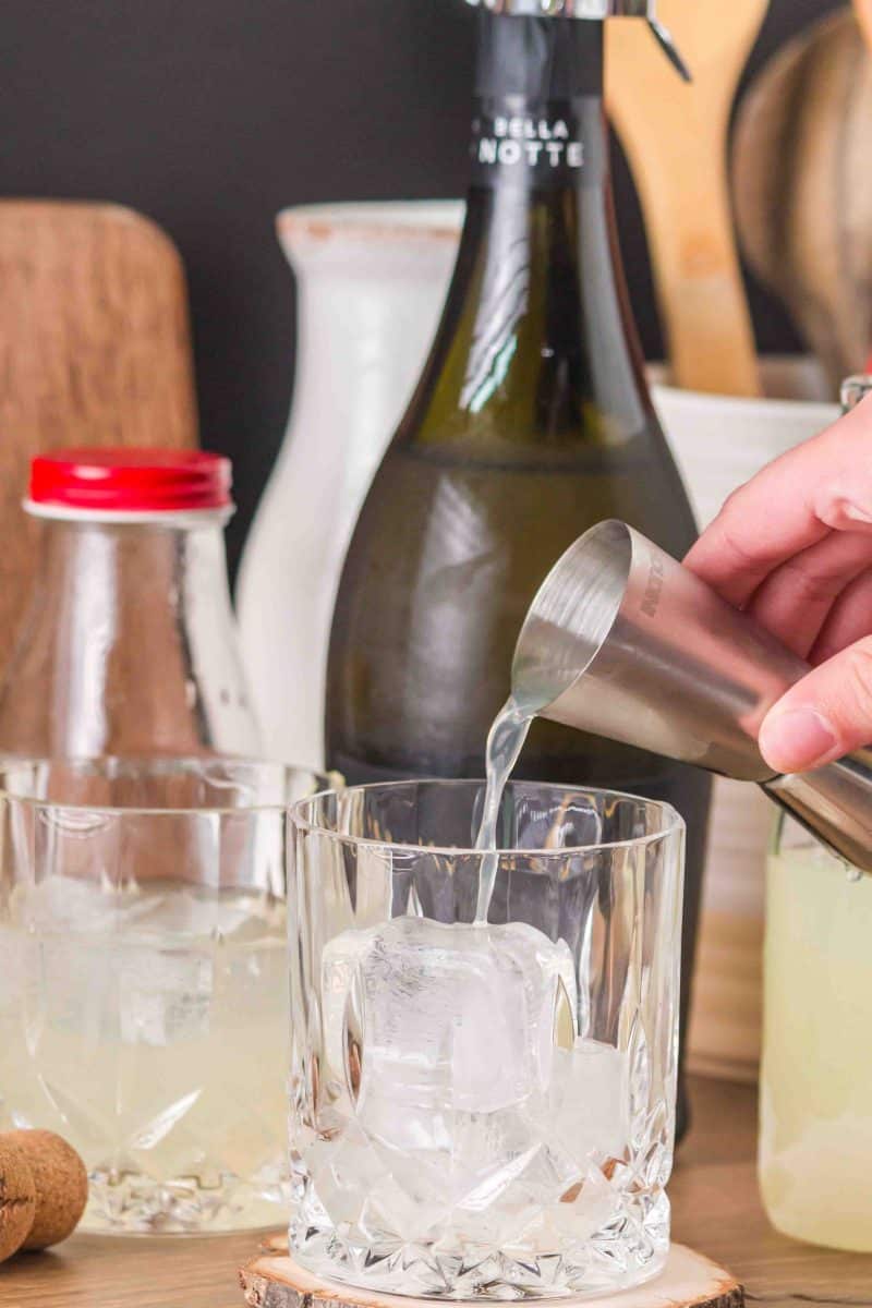 Sideview of a rocks glass with a large ice cube. A hand is pouring Limoncello liqueur over the ice cube. Hostess At Heart