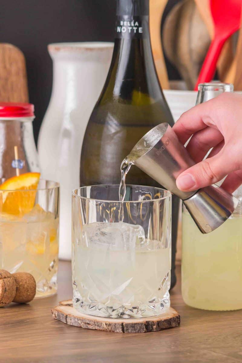 A rocks glass pouring Prosecco over an ice cube, limoncello liqueur, and club soda to make an Italian cocktail - Hostess At Heart