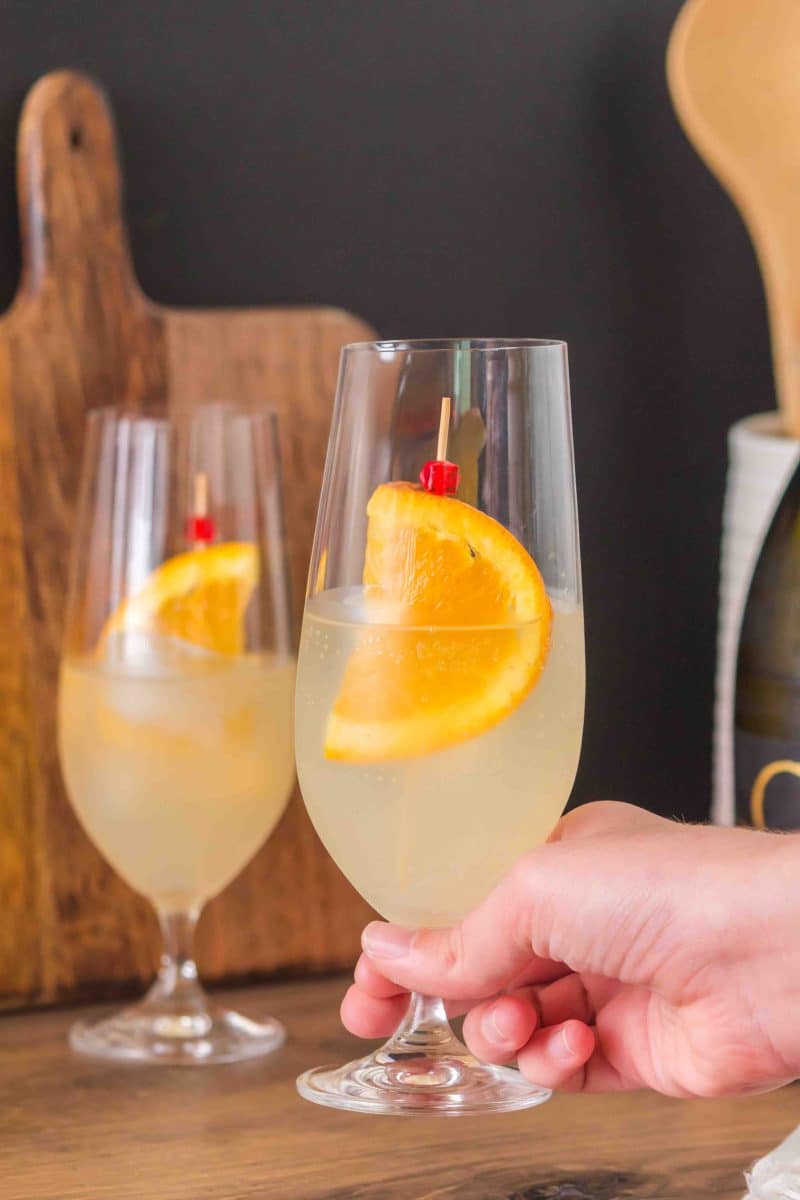 A hand holding a finished Limoncello cocktail in a champagne glass garnished with a orange wheel, showing the pale yellow bubbly drink - Hostess At Heart