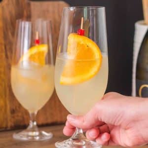 Finished Limoncello Spritz cocktail in a champagne glass garnished with a orange wheel and, showing the pale yellow bubbly drink - Hostess At Heart