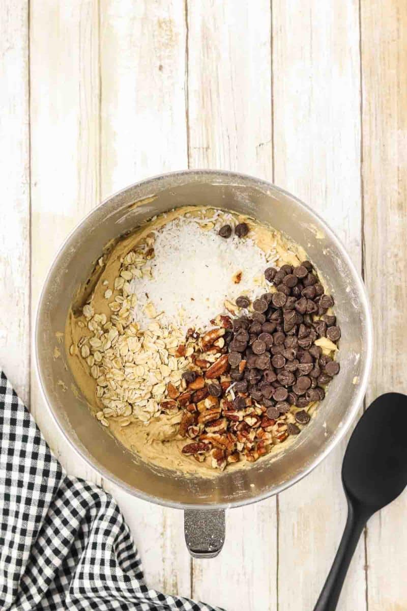 Mixins including shredded coconut, chopped pecans and old-fashioned oats into thick beige cookie dough in large medal mixing bowl, ingredients partially mixed showing distinct layers. Hostess At Heart
