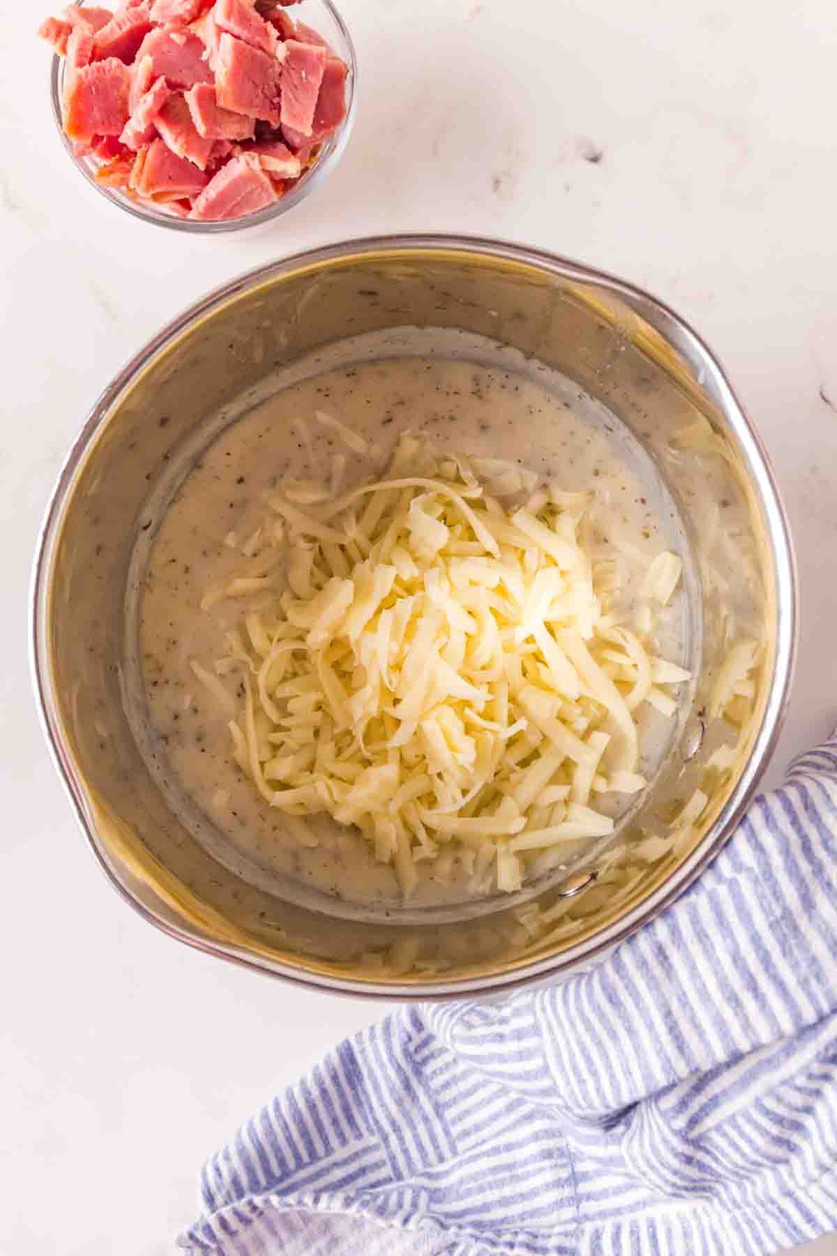 Shredded mozzarella cheese being stirred into creamy white sauce in saucepan - Hostess At Heart