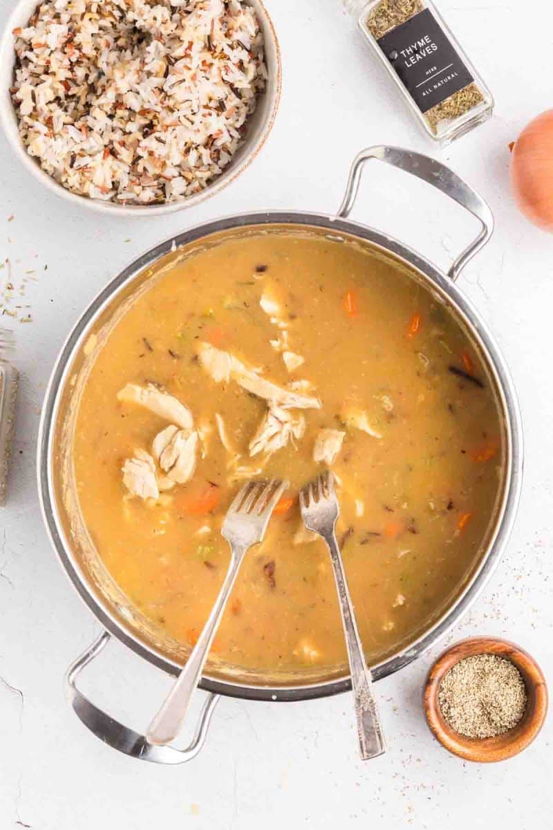 Shredded chicken tenders cooking in simmering broth with herbs and carrots visible for homemade chicken and wild rice soup. Hostess At Heart