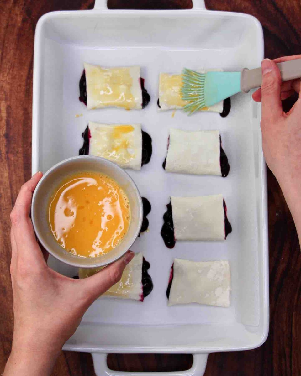 A pastry brush applying egg wash to blueberry puff pastry rolls in a baking dish - Hostess At Heart