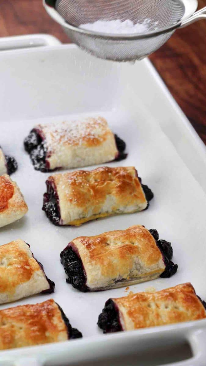 A sideview of a baking dish filled with golden brown blueberry pastries being dusted with powdered sugar - Hostess At Heart