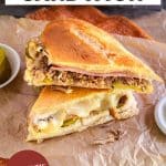 A authentic pressed Cuban Sandwich cut in half on parchment paper oozing with melted cheese over pork and ham. Hostess At Heart