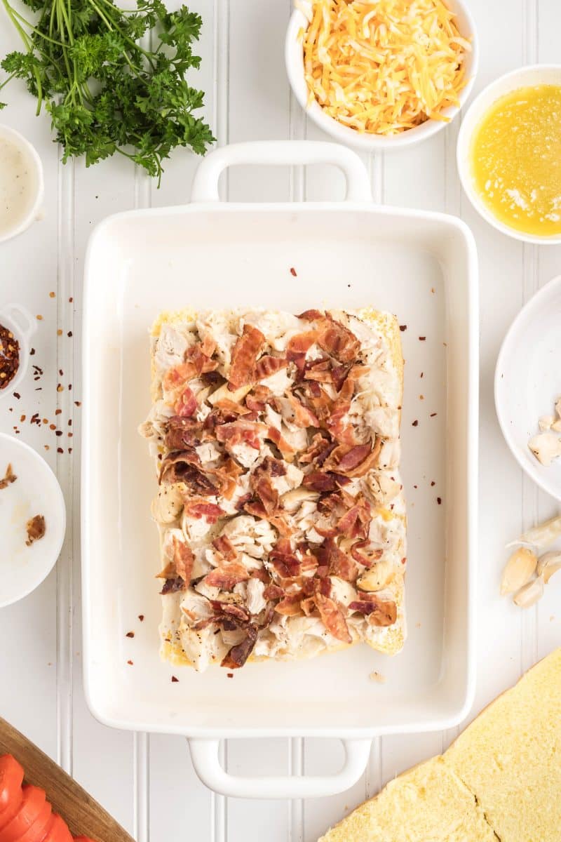 Slider rolls being assembled with layers of shredded chicken and crumbled bacon pieces evenly distributed across rolls. Hostess At Heart