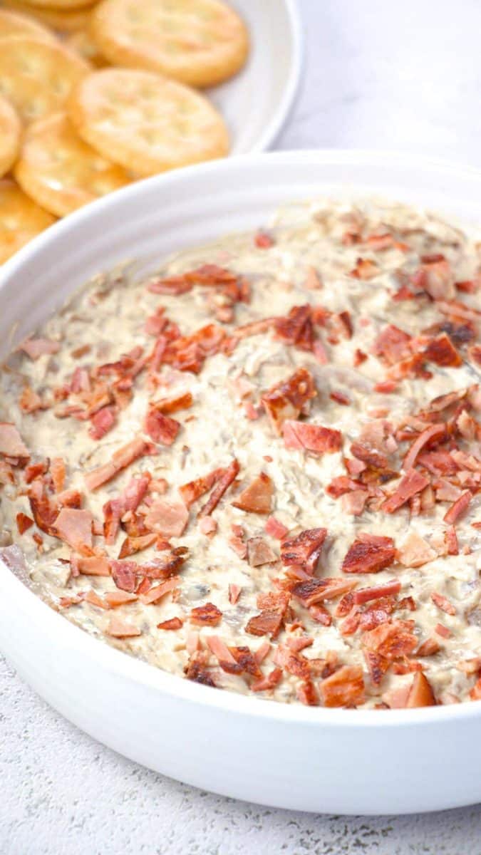 Golden creamy caramelized onion bacon dip topped with crispy bacon pieces in white serving bowl. Hostess At Heart