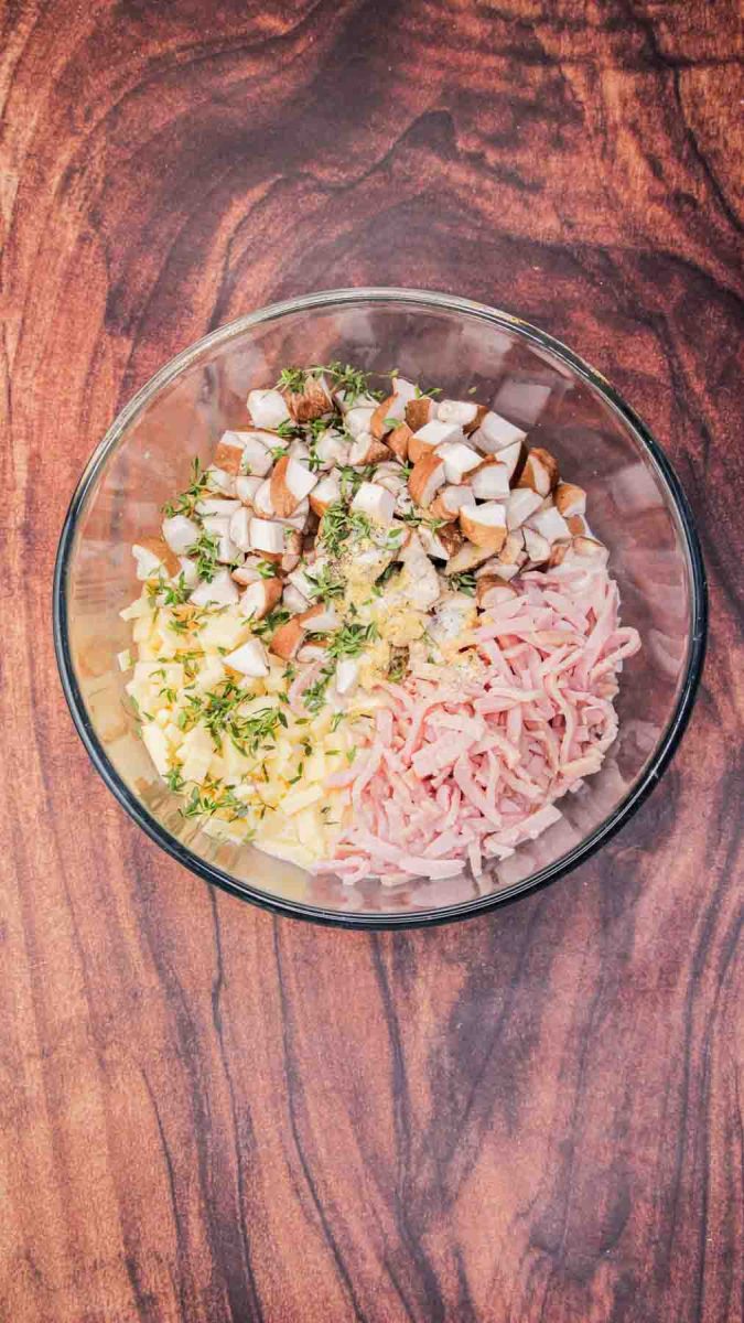 Ingredients for breakfast strata including, ham, mushrooms, eggs, milk, and shredded cheese in a mixing bowl on kitchen counter preparing to be stirred together. Hostess At Heart