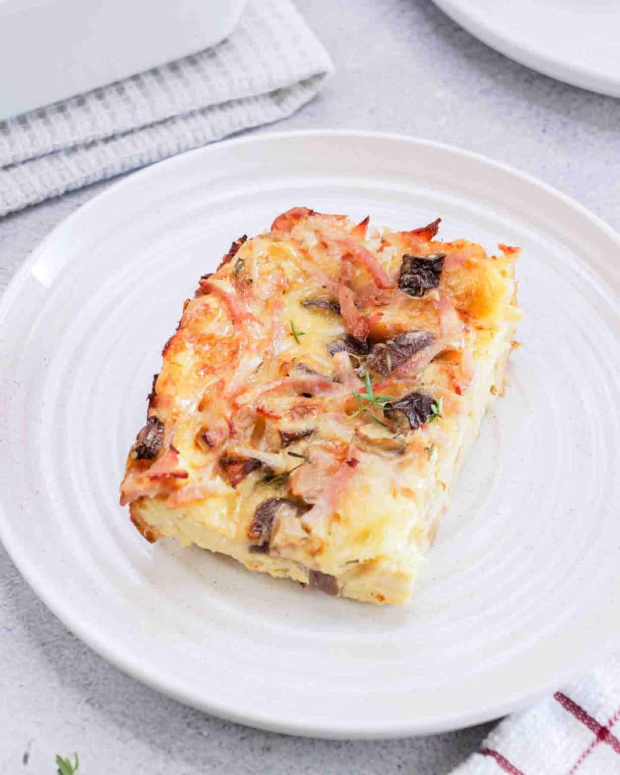 Close up of breakfast strata slice loaded with custardy bread, melted cheese, ham and mushrooms on a white plate.