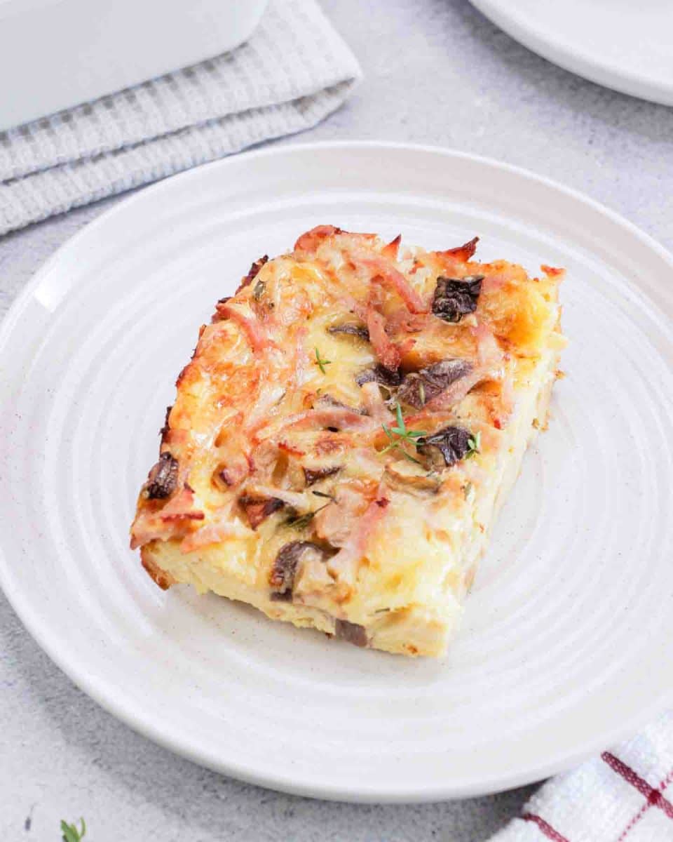 Close up of breakfast strata slice loaded with custardy bread, melted cheese, ham and mushrooms on a white plate.