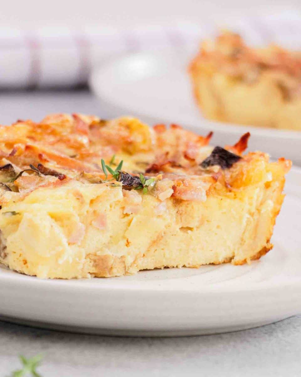 Close up of breakfast strata slice showing layers of custardy bread, melted cheese, ham and mushrooms. Hostess At Heart