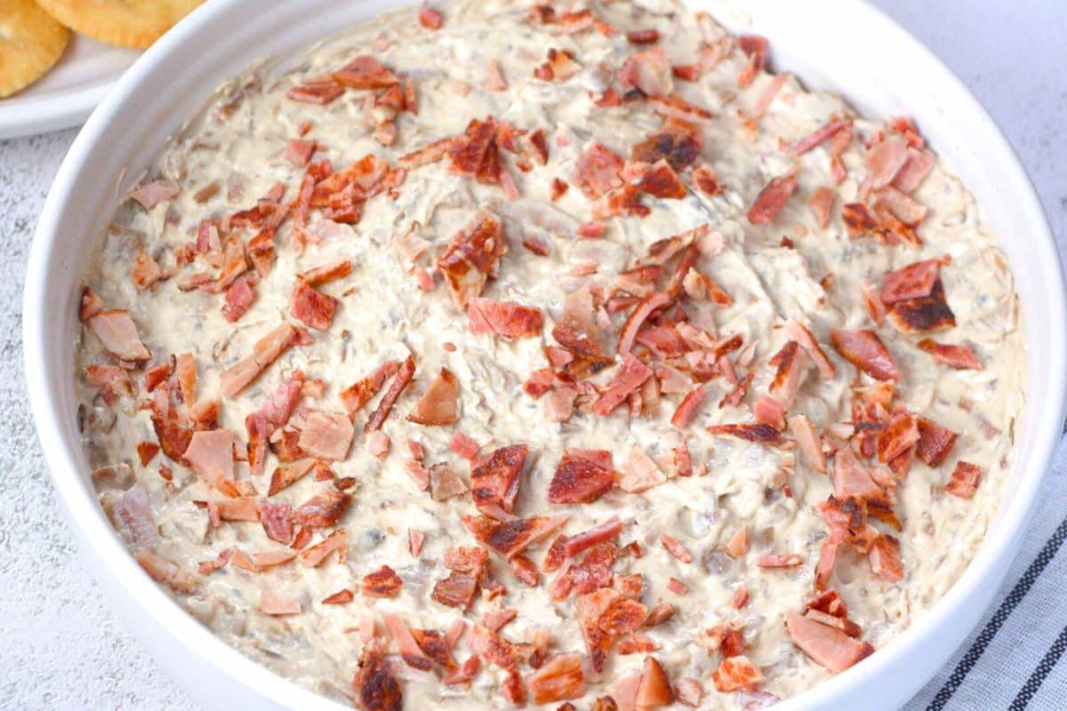 Golden creamy caramelized onion bacon dip topped with crispy bacon pieces in white serving bowl. Hostess At Heart