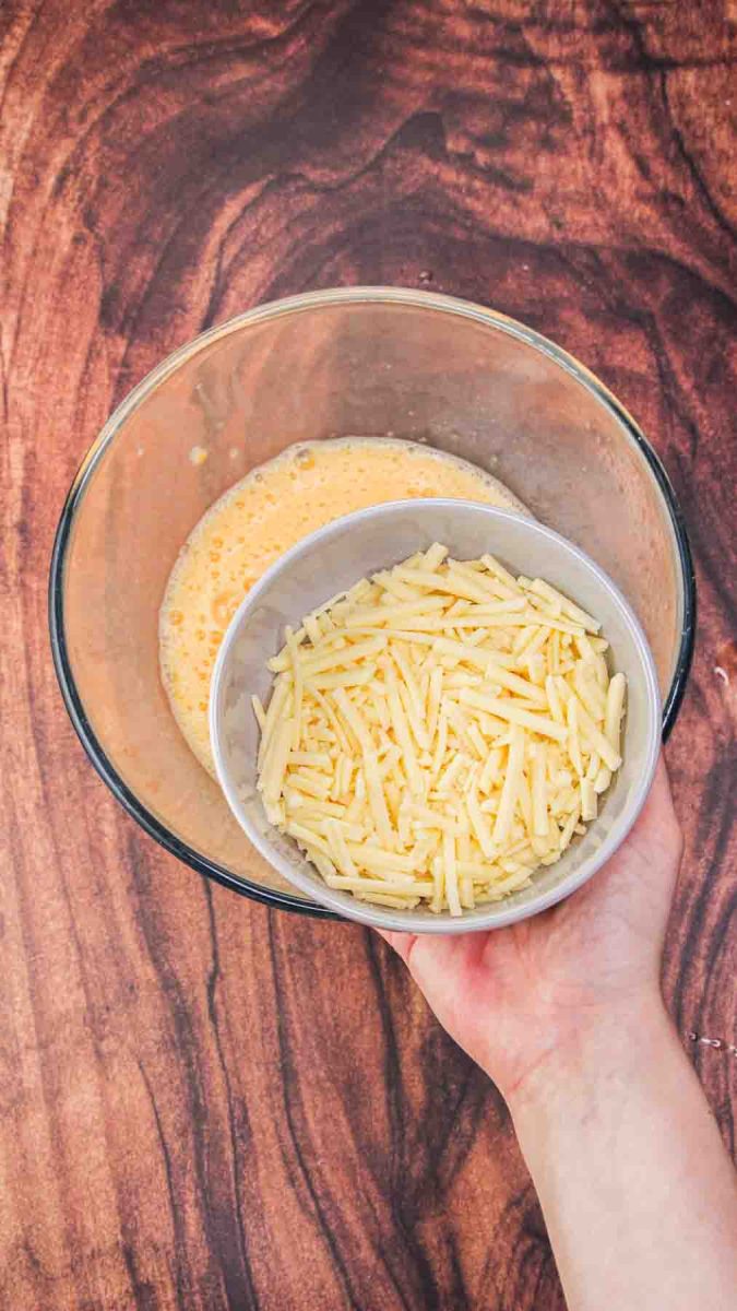 A bowl of shredded cheese being added to an egg mixture to make a Ham and Cheese Mushroom Breakfast Strata. Hostess At Heart