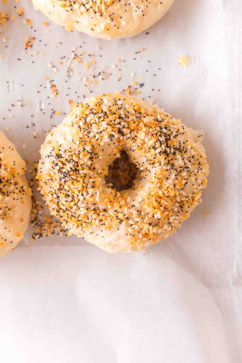 Top down view of a boiled breakfast bagel garnished with everything bagel seasoning on a sheet of parchment paper ready for baking - Hostess At Heart