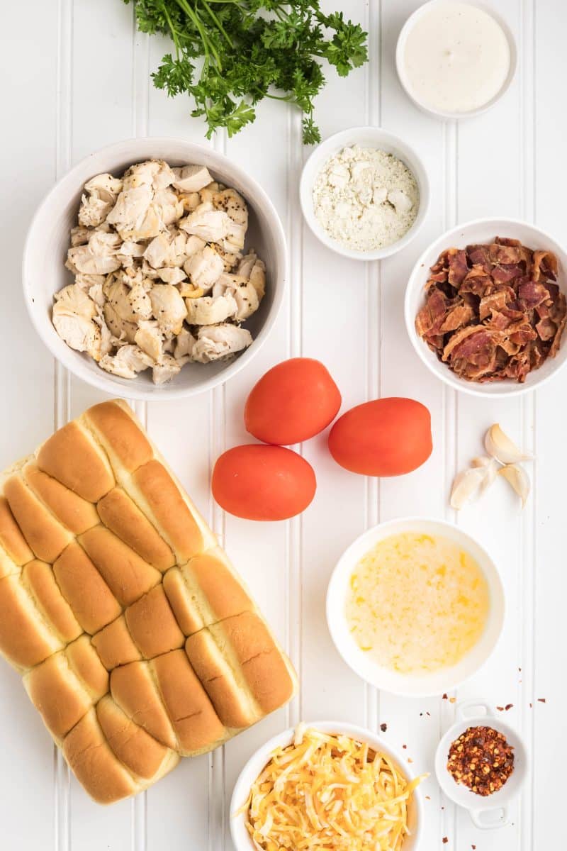 Ingredients for chicken bacon ranch sliders arranged on counter including Hawaiian rolls, chopped chicken, crispy bacon, ranch dressing, shredded cheese, red chili flakes, and fresh tomatoes. Hostess At Heart
