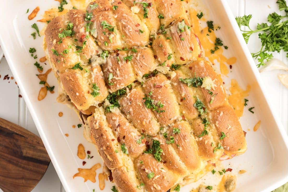 Top down view of baked chicken bacon ranch sliders showing layers of melted cheese, bacon, chicken, and tomato with visible garlic butter and a fresh parsely garnish on a golden top bun. Hostess At Heart