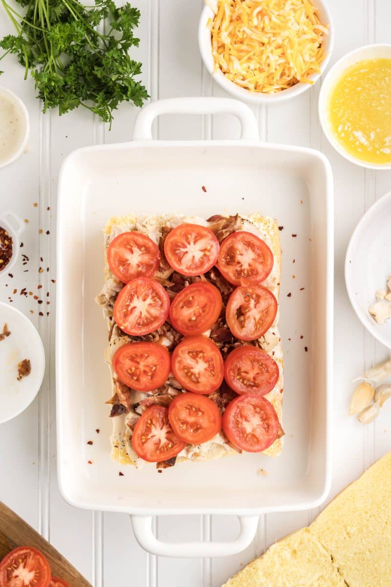 Chicken Slider rolls being assembled with layers of chopped chicken and crumbled bacon pieces evenly distributed across rolls and topped with slices of fresh tomatoes. Hostess At Heart