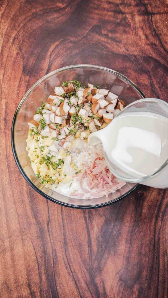 Pouring milk into a mixing bowl filled with breakfast strata ingredients including, ham, mushrooms, eggs, milk, and shredded cheese for make ahead breakfast casserole. Hostess At Heart